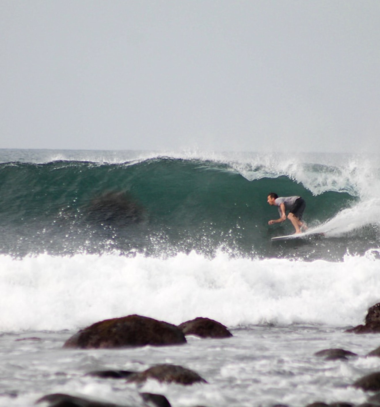 A surfer riding the face of a green wave, with rocks and whitewater in the foreground.